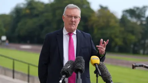 Mike Nesbitt standing behind four microphones while speaking to the media. He is wearing a dark suit with a white shirt and a pink tie. 