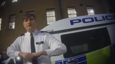 A hidden camera shot, taken slightly from below and at an angle, of an officer in police uniform, a white man with brown hair in a side-parting and a with a cleft chin, standing in front of a police van in the courtyard of Charing Cross police station, with windows of the station all around. He is pulling with his right hand on the fingers of his left as he discusses taking fingerprints by force