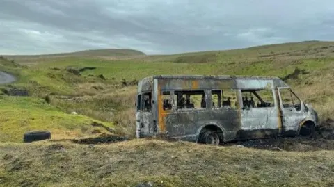 St Joseph's R.C. Primary School A torched school bus on a bank with green hills seen behind it. A tyre can be seen behind the vehicle, which is blackened with no wheels. A curving road can just be seen out of shot on the left hand side. 
