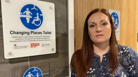 Felicity Benyon in front of a Changing Places toilet. A white sign with a blue logo indicates the toilet is for people with disabilities or additional needs.