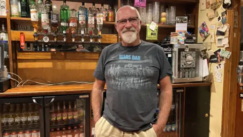 A man wearing a t-shirt that says 'Mars Bar', stood behind a series of drink cabinets full of bottles of beer. 