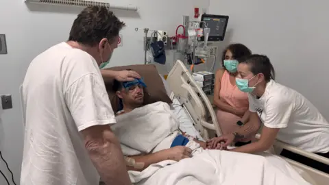 Handout A man lies in a hospital bed, covered with a white blanket. His face is very bruised and there are a number of drips and monitors visible in the background. A man holds a compress to his head. A woman is bending down to look at him, holding one of his hands. 