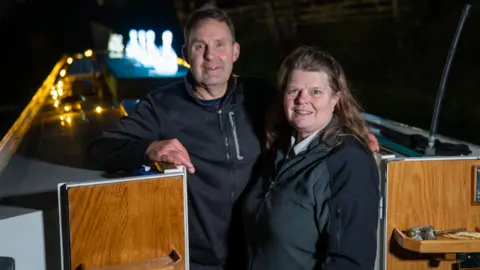Photograph of Samantha and Jim Atkins on board their boat. They both have blue eyes and brown hair and smile at the camera. Jim stands behind samantha, wearing a black jacket and has his left arm leaning on the top of his boat. Samantha wears a black headband and grey and black jacket, has brown shoulder left hair and smiles with her teeth. the top of their boat can be seen behind them. Four swan lanterns line the centre of the boat. 