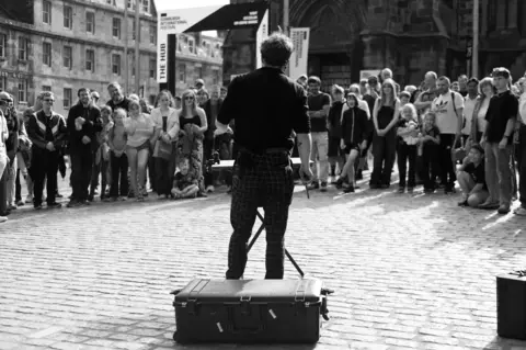 Chris Foote/BBC Scotland A black and white image taken from behind a street artist as he performs to a circle of people on a cobbled square in Edinburgh. A suitcase sits behind him on the ground, a table in front where he seems to be performing a trick of some kind