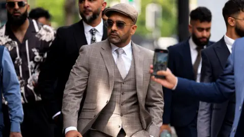 A man in a beige three-piece suit and a beige cap walks outside, surrounded by other men in suits. The man is wearing sunglasses and he has a neatly trimmed black beard.
