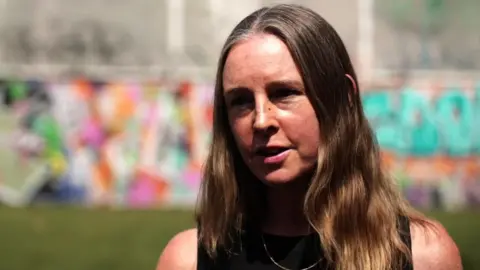 Tara Darby in a black vest top with long hair standing on the sports pitch.