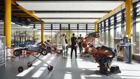 Riba Four students stand in a large room with floor to ceiling windows and yellow columns. A small car and a robotic arm are on display.