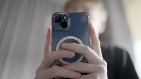 A close-up image of a teenage boy holding an iPhone with both hands. The phone and its camera lenses dominate the image, while the boys face is hidden and out of focus