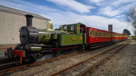A green steam railway engine pulling red carriages on the railway line in Port Erin with blue sky above.