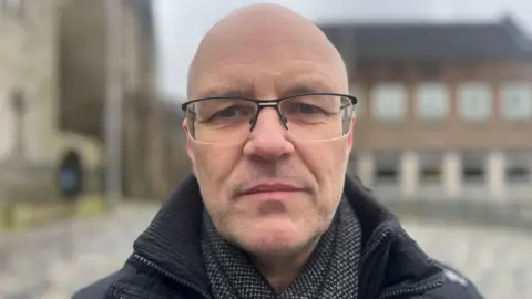 Dominic is looking directly at the camera. He is standing outside on the street and in the back left of the image is a church building. Dominic has a bald head and is wearing glasses.