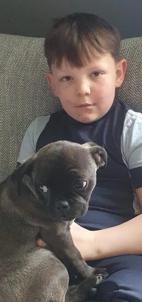 Family handout Rory when he was younger. In it, a boy aged about eight year old is sat on the sofa with a dark-coloured pug dog. He is wearing a grey T-shirt with a dark blue vest over the top. He is not smiling but is looking into the camera. He has dark hair.