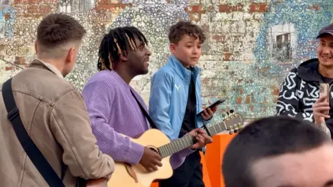 Danny Fullbrook/BBC A boy wearing a light blue jacket is stood next to Myles who is singing and playing guitar next to him