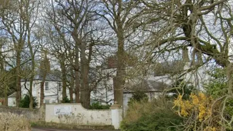 Google Howlish Hall care home is a white building tucked behind a row of trees and a white wall.
