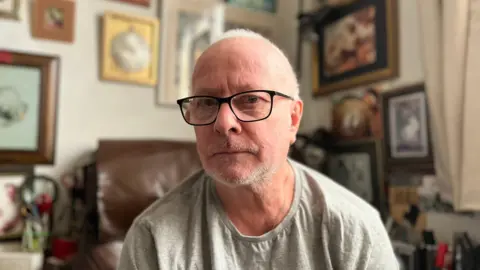 Aidan Zeall wearing a grey t shirt and glasses in his living room with pictures behind him. he has grey hair