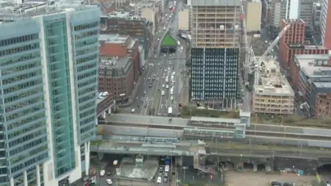 Aerial shot of city from a drone. There are lanes of vehicles and a bridge over them. There are also tall buildings and a number of smaller buildings and a crane in the picture. 