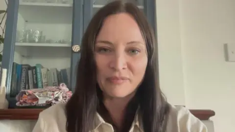 Alex sits on a video call. She is wearing a white shirt and has shoulder-length black her. Behind her is a mantlepiece with books and glasses on show.