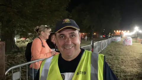 David Hesketh with short dark hair wearing a blue cap and yellow hi vis next to a metal barrier. A few spectators are visible.  It is dark but lights are visible in the background