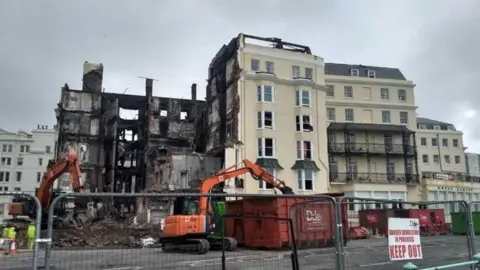 A large Regency-style building. The left of the building has been destroyed by fire and demolition vehicles are tearing it down