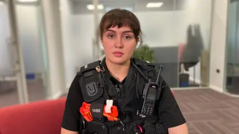 A female police officer in uniform. She is standing indoors looking at the camera.