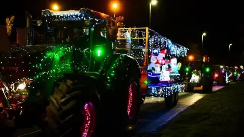 There are a number of tractors lit up with Christmas lights. The tractor is green. 