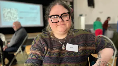 BBC/Richard Moss Brittania Douglas has dark-rimmed large glasses and some tattoos on her forearms. She is smiling and wears a patterned jumper.