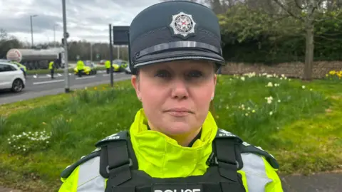 A woman wearing a PSNI uniform looks at the camera. She is wearing a dark-green hat and a fluorescent yellow jacket. Behind her is a patch of grass, and further in the distance is a road on which several other police officers are standing. 