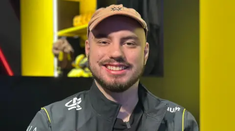 Derke wears a grey zip up fleece with a white logo on it. He wears a brown cap and has a short brown beard
