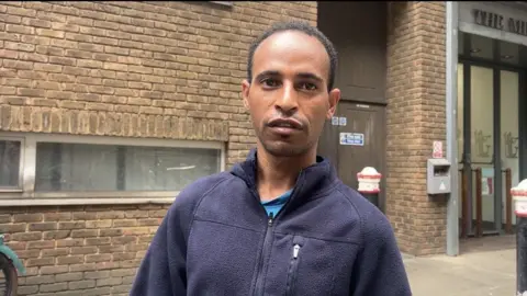 A man is wearing a dark blue fleece and standing in front of a brick wall