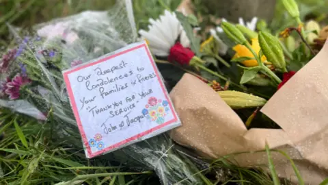 Two bouquets of flowers left at the scene of a helicopter crash. The flowers are on a grass verge. A card on one of the bunches reads: "Our deepest condolences to your families and friends. Thank you for your service. John and Jacqui.