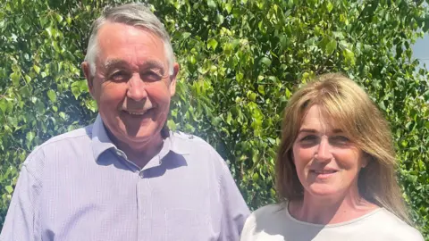 The Souttars in blue and white shirts standing side by side smiling in front of a tree