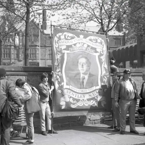 Alan Van Wijgerden A black and white image of two men holding up a Bentley National Union of Miners banner with an image of Arthur Scargill on it. Other people are standing around them. 