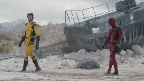 Disney An image from the new deadpool and wolverine film. Jackman is on the left in his yellow wolverine outfir, whilst Ryan Reynolds is on the right in his red and black deadpool costume. In the background there is an old ship and it is some sort of dust plain.