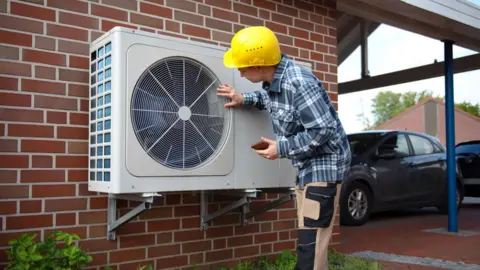 An engineer in a checked shirt and yellow safety helmet examines a heat pump on the side of a house.
