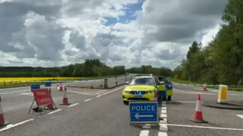 A police car sits among road closure signs and orange and red traffic cones at an A-road junction following a crash in the Cotswolds. The passenger door of the police car is open as a police officer reaches inside. It is a partly cloudy spring day and a field on the opposite side of the road is full of yellow flowers.