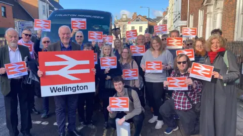 Supplied A group of people, standing next to a minibus, holding railway station signs saying "Wellington"