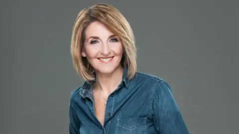 BBC Scotland Kaye Adams smiling straight at the camera. She is wearing a blue shirt and is standing against a grey background.
