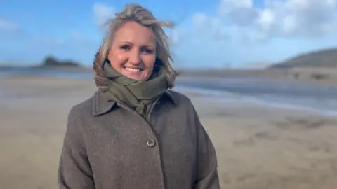 A woman wearing a coat and scarf smiles on a beach 