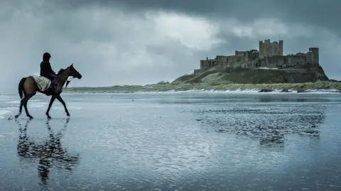 Jim Scott Photography A person riding a horse on a water covered sandy beach. They are heading toward a grand stone castle on grassy mound in the distance. The sky overhead is grey and cloudy, giving a dramatic quality to the scene.