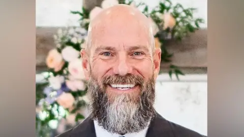 Supplied A head and shoulders image of a bald man with a beard. He is smiling and looking into the camera. Behind him are some flowers.