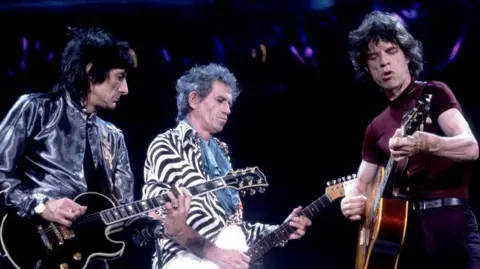 Getty Images Three men on stage performing and playing guitars. The man on the left has dark black hair and wears a shiny metallic grey jacket. The middle man has black and grey hair and wears a stripey black and white jacket. The man on the right has long dark hair and wears a burgundy T-shirt and black trousers. 