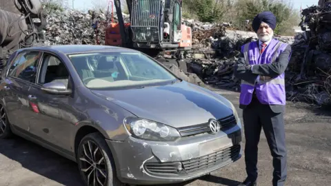 City of Wolverhampton Council A man in a purple turban and purple high-vis jacket over a suit is standing next to a silver VW Golf in the middle of a scrapyard