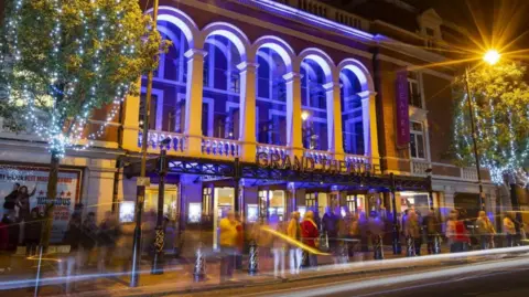 Jonathan Hipkiss The theatre is lit up at night with people walking by outside. It has a purple hue from the lighting  with two trees either side with lights on.