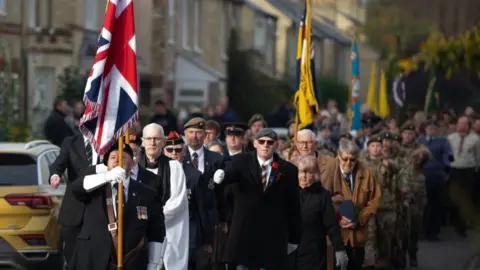 John Dalston Parade led by the union flag carried by an RBL member with a priest alongside. Other standards are visible in the background