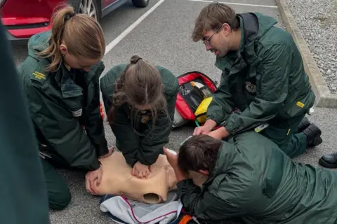 Lorna Edwards A group of paramedic students practicing CPR during training. The students are kneeled on a concrete floor outside, leaning over a plastic dummy, checking airwaves and conducting CPR. The students are wearing a dark green paramedic uniform.