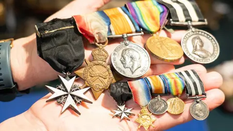 A person holding five medals with multi-coloured ribbons alongside five matching miniature dress medals.