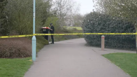 Jamie Niblock/BBC Yellow police tape attached to a lamppost, it is cordoning off an area of a park. A police officer is behind the tape and appears to be writing something down.