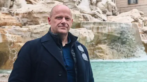 Facebook Grant Paterson, who has balding shaved hair and is wearing a dark blue shirt with a navy blue woollen coat, stands at the Trevi Fountain in Rome. 