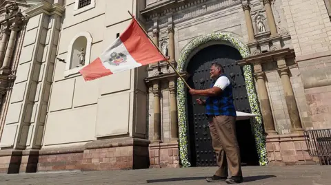 Getty Images A man wearing brown trousers and a black and blue plaid shirt waves a Peruvian flag in front of the Metropolitan Cathedral in Lima after the election of Pope Leo XIV on 8 May 2025