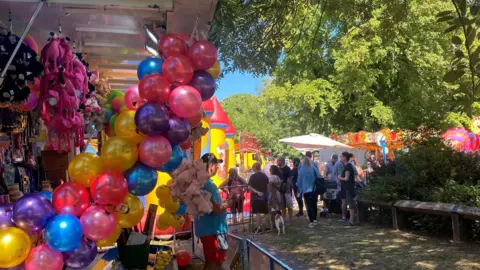 Christine Brant A hook-a-duck stand in a park. It has toys and balloons on display. People are gathering in the distance.