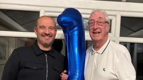 Two men one old and one younger smiling in a conservatory. They are holding a large helium birthday balloon.